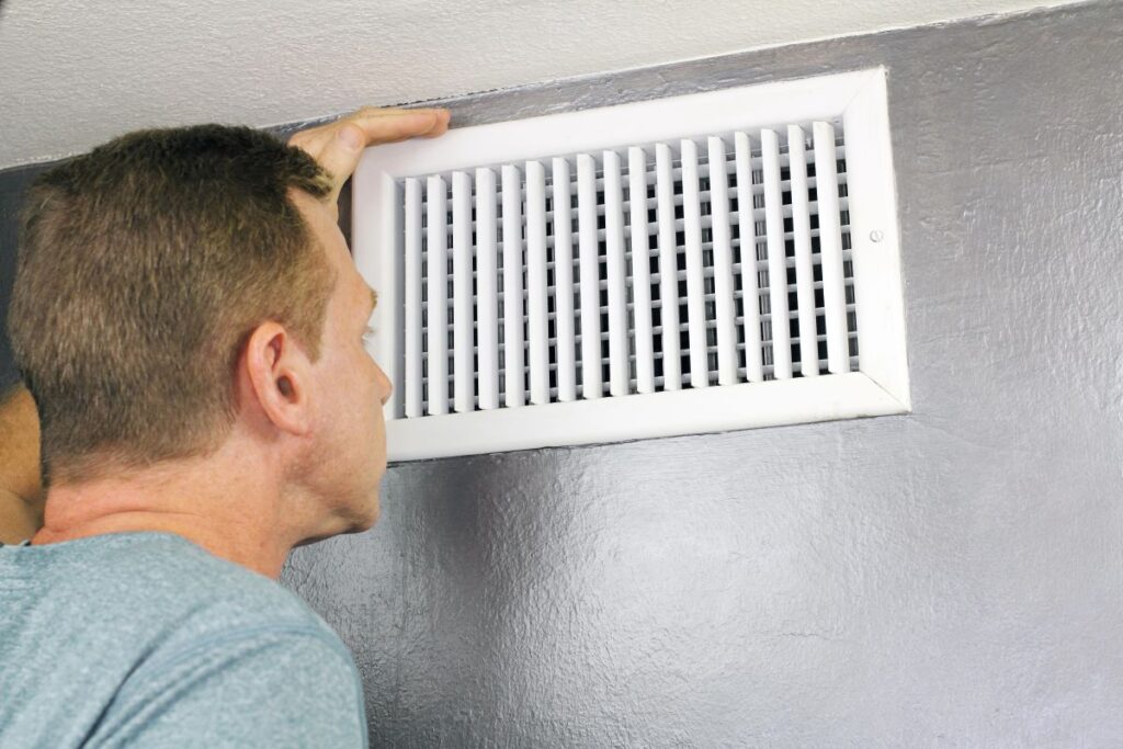 A man inspecting an HVAC vent on a wall, likely trying to diagnose common HVAC smells.