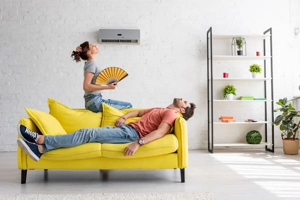 A couple in a hot room under an air conditioning looking for tips for running an air conditioning in extreme heat.