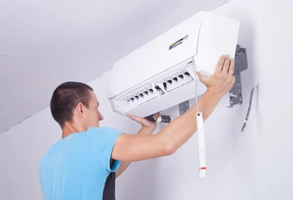 A man fixing an air conditioner inside a living room, showing the maintenance of an AC that is not working in hot weather.