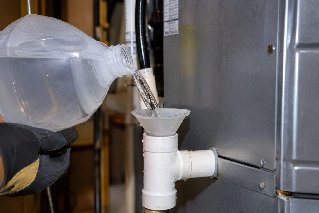 A homeowner pours cleaning solution into a PVC pipe funnel to clear an ac drain line clogged on an air handler.
